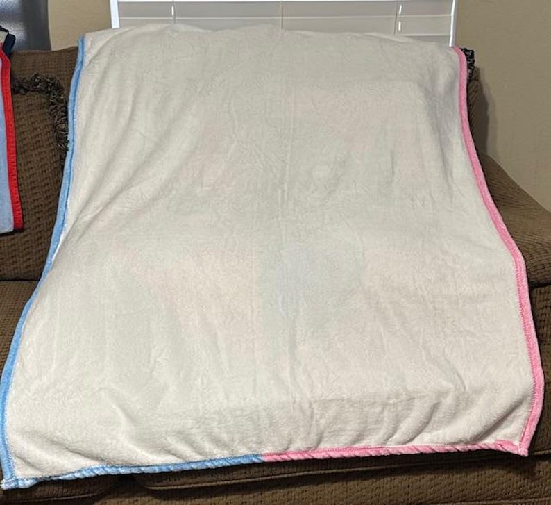 Może przedstawiać: Miękki, biały koc z niebieską i r&oacute;żową obw&oacute;dką jest narzucony na brązową sofę. Koc ma miękką, pluszową fakturę i wydaje się być wykonany z przytulnego materiału. Krawędzie są starannie wykończone kontrastową lam&oacute;wką.