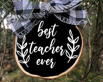 May include: A black wooden ornament with the text "Best Teacher Ever" in white lettering. The ornament is decorated with a white and black checkered bow and a small sprig of leaves on each side of the text.