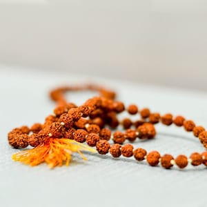 Puede incluir: Un collar de cuentas mala marrón con un borla de hilos naranja y amarillo. Las cuentas están hechas de materiales naturales y están enhebradas en un cordón delgado.