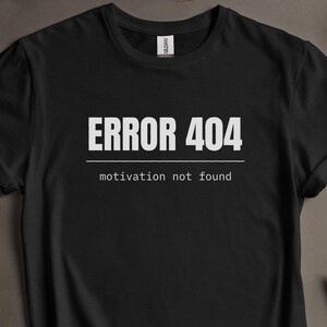 May include: A black short-sleeved t-shirt featuring the white text ERROR 404 above a horizontal line and the text motivation not found below it. The shirt has a crew neckline and a white brand label visible at the inside of the collar.