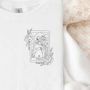 May include: White crew neck sweatshirt with a black and white Virgo tarot card design. The design features a woman, flowers, and the word "Virgo". The sweatshirt is displayed with a beige hat and dried palm leaves.