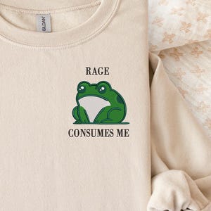 Pu&ograve; includere: Una felpa girocollo color sabbia con un ricamo raffigurante una rana verde sul petto sinistro. Sopra la rana, il testo recita RAGE, e sotto la rana, il testo recita CONSUMES ME. La felpa presenta maniche lunghe e un colletto a coste.