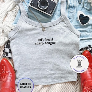 May include: A casual fashion flat lay featuring a gray athletic heather tank top with the text "soft heart sharp tongue", light wash denim shorts, heart-shaped sunglasses, a vintage camera, and red cowboy boots. A necklace and a black and white checkered scarf complete the look.