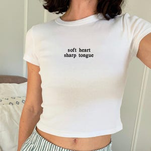 May include: White, short-sleeved crop top with the text "soft heart sharp tongue" in black. The top is paired with green and white striped pajama shorts. The image is taken indoors.