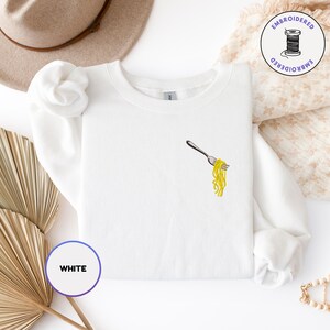 May include: White sweatshirt with an embroidered fork holding yellow spaghetti. The design is on the left chest. The word "WHITE" is in a circle in the lower left corner. A hat and decorative items are in the background.
