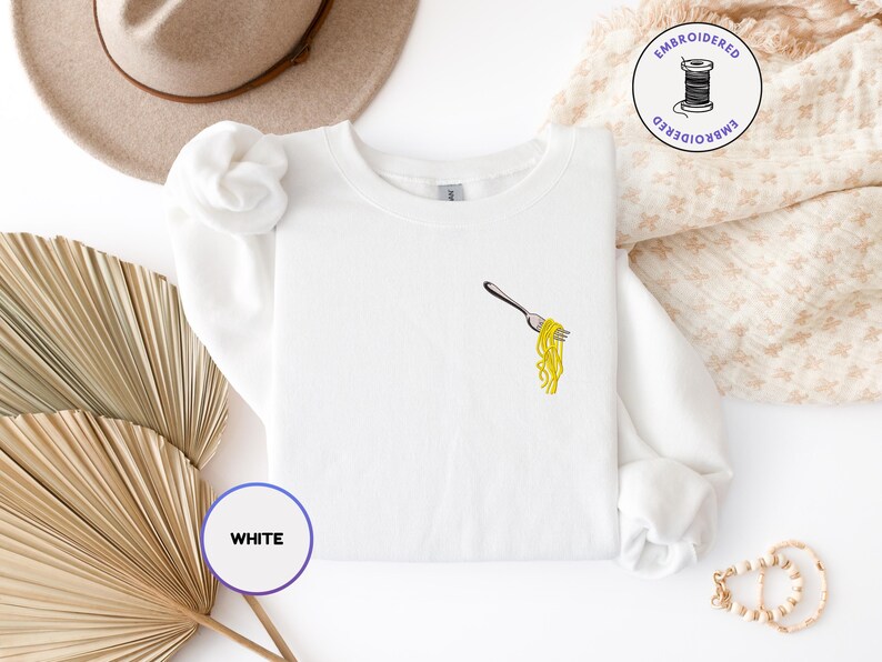 May include: White sweatshirt with an embroidered fork holding yellow spaghetti. The design is on the left chest. The word "WHITE" is in a circle in the lower left corner. A hat and decorative items are in the background.