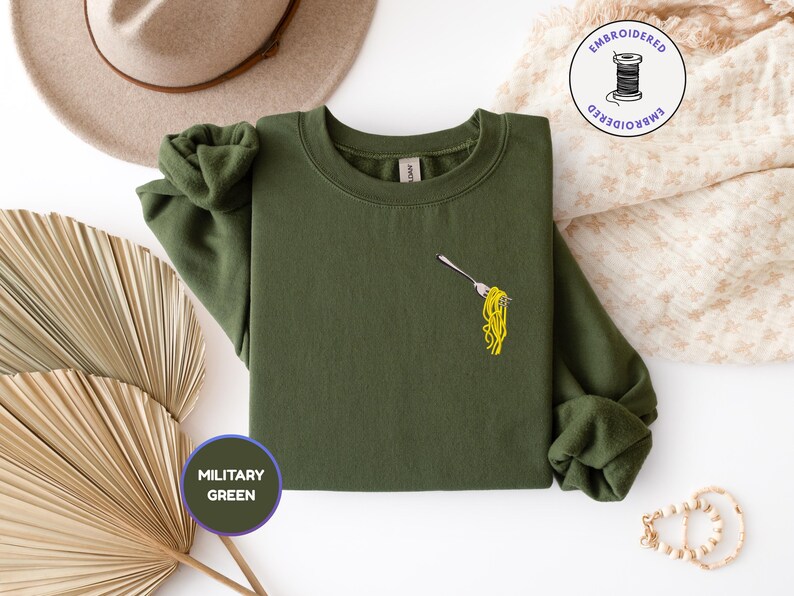 May include: Military green sweatshirt with an embroidered fork holding yellow spaghetti. The sweatshirt has a crew neck and long sleeves. The image also includes a hat, a decorative fan, and a small embroidered logo.