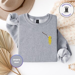May include: A light gray sweatshirt with a crew neck and long sleeves. The sweatshirt features an embroidered design of a fork with yellow spaghetti. The words "SPORTS GRAY" are printed on a purple circle. A tan hat and decorative items are in the background.