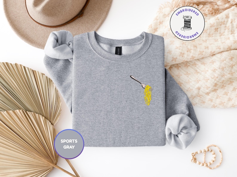 May include: A light gray sweatshirt with a crew neck and long sleeves. The sweatshirt features an embroidered design of a fork with yellow spaghetti. The words "SPORTS GRAY" are printed on a purple circle. A tan hat and decorative items are in the background.