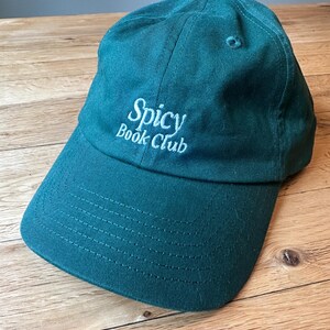 K&ouml;nnte beinhalten: T&uuml;rkisfarbene Baseballkappe mit dem gestickten Text "Spicy Book Club" in Wei&szlig;. Die Kappe hat einen gebogenen Schirm und einen Knopf oben. Der Hut ist aus weichem Stoff und liegt auf einer Holzoberfl&auml;che.
