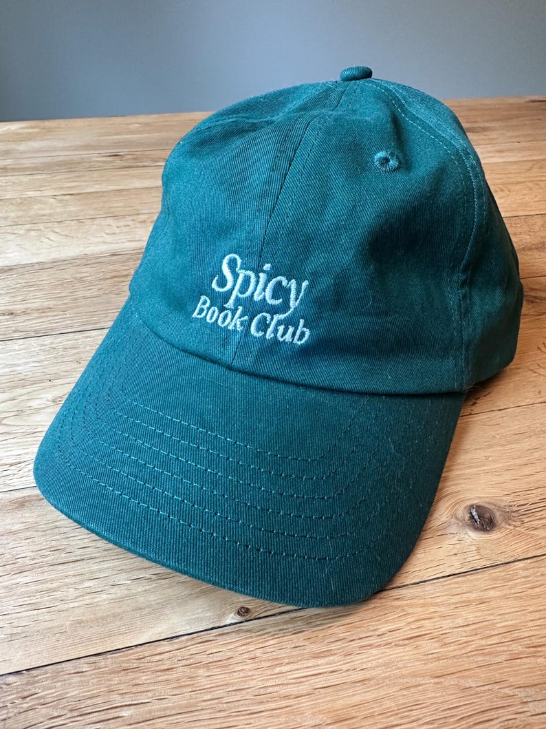 K&ouml;nnte beinhalten: T&uuml;rkisfarbene Baseballkappe mit dem gestickten Text "Spicy Book Club" in Wei&szlig;. Die Kappe hat einen gebogenen Schirm und einen Knopf oben. Der Hut ist aus weichem Stoff und liegt auf einer Holzoberfl&auml;che.