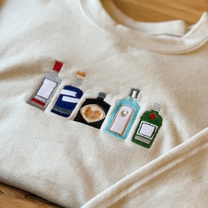 Op de afbeelding: Een wit sweatshirt met een geborduurd ontwerp van zes verschillende flessen alcohol. De flessen staan in een rij en zijn allemaal in verschillende kleuren en vormen.