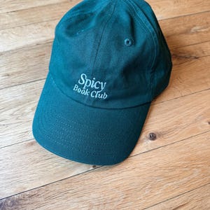 K&ouml;nnte beinhalten: Eine tealfarbene Baseballkappe mit dem Text "Spicy Book Club" in Wei&szlig; gestickt. Die Kappe hat einen gebogenen Schirm und einen kleinen Knopf oben. Der Hut ist aus einem weichen Stoff gefertigt.