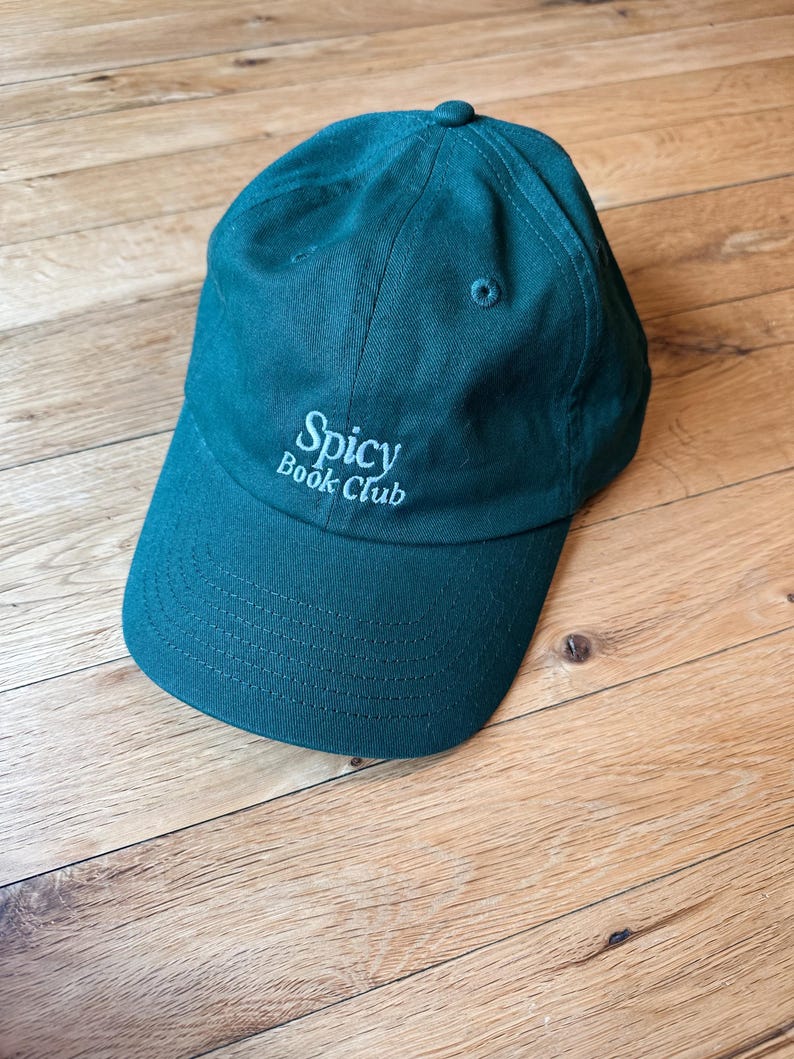 K&ouml;nnte beinhalten: Eine tealfarbene Baseballkappe mit dem Text "Spicy Book Club" in Wei&szlig; gestickt. Die Kappe hat einen gebogenen Schirm und einen kleinen Knopf oben. Der Hut ist aus einem weichen Stoff gefertigt.