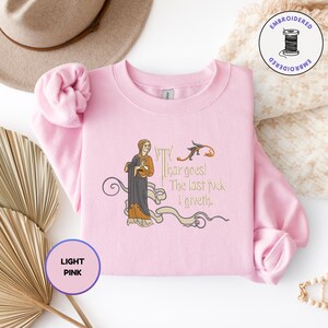 Puede incluir: Una sudadera de cuello redondo rosa claro con un bordado de estilo medieval que muestra a una persona con una t&uacute;nica oscura y el texto 'Thar goes! The last fuck I giveth.' El dise&ntilde;o incluye adornos decorativos en tonos dorados y marrones.