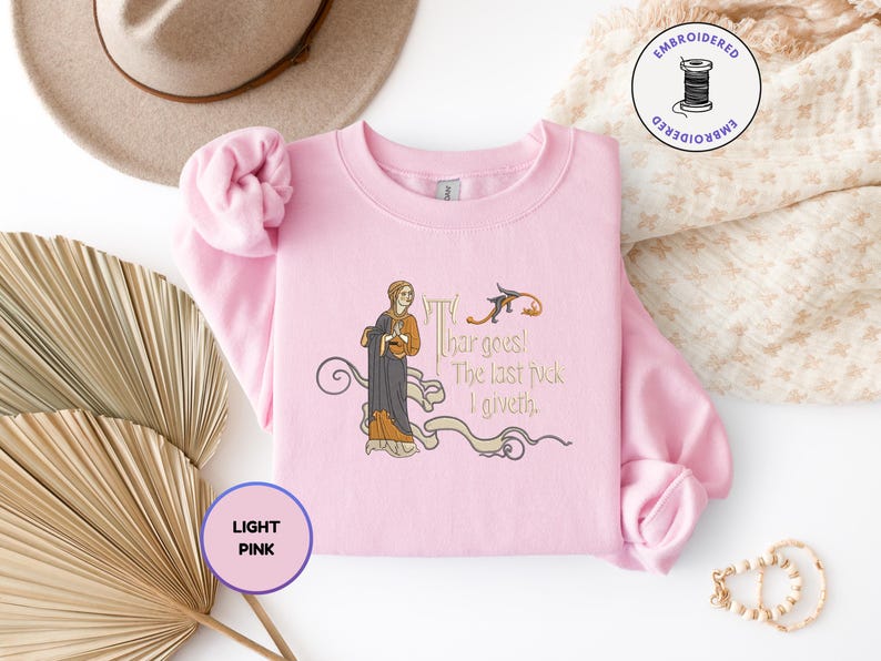 Puede incluir: Una sudadera de cuello redondo rosa claro con un bordado de estilo medieval que muestra a una persona con una t&uacute;nica oscura y el texto 'Thar goes! The last fuck I giveth.' El dise&ntilde;o incluye adornos decorativos en tonos dorados y marrones.