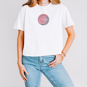May include: A white short-sleeved t-shirt featuring a circular graphic on the chest. The design includes the word SCORPIO in dark pink letters, a pink scorpion illustration, blue wave patterns, and small star shapes on a light pink background.