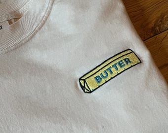 May include: A white t-shirt featuring an embroidered patch on the chest. The patch is shaped like a stick of butter, colored yellow with blue text that reads BUTTER, outlined in black thread. The shirt has a ribbed crew neckline.