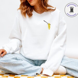 May include: White sweatshirt with an embroidered design of a fork holding yellow spaghetti. The sweatshirt has a crew neck and long sleeves. The image also includes the text "EMBROIDERED". The model is wearing light blue jeans.
