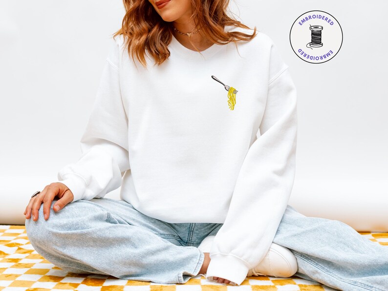 May include: White sweatshirt with an embroidered design of a fork holding yellow spaghetti. The sweatshirt has a crew neck and long sleeves. The image also includes the text "EMBROIDERED". The model is wearing light blue jeans.