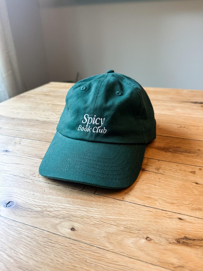 K&ouml;nnte beinhalten: Dunkelgr&uuml;ne Baseballkappe mit dem wei&szlig;en, gestickten Schriftzug "Spicy Book Club". Die Kappe hat einen gebogenen Schirm und ein klassisches Sechs-Panel-Design, ideal f&uuml;r den Alltag.