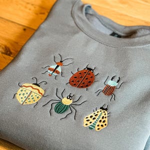 May include: A grey crewneck sweatshirt featuring an embroidered design of six distinct insects. The embroidery includes a ladybird, a spider, and various beetles in shades of red, yellow, green, brown, and light blue, all stitched with black thread.
