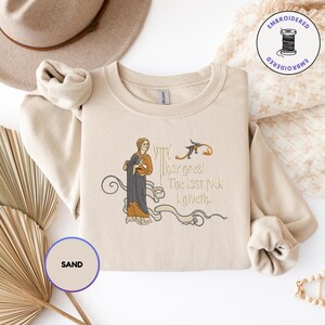 Puede incluir: Una sudadera de cuello redondo color arena con un bordado que muestra una figura de estilo medieval con t&uacute;nica oscura y el texto 'Thar goes! The last fuck I giveth.' El dise&ntilde;o incluye l&iacute;neas curvas decorativas y una peque&ntilde;a criatura.