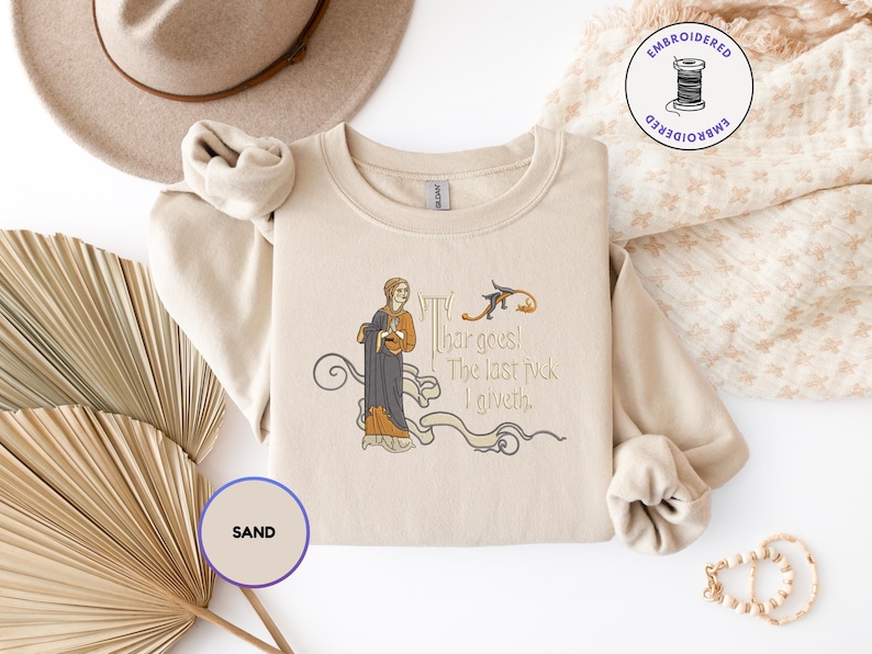Puede incluir: Una sudadera de cuello redondo color arena con un bordado que muestra una figura de estilo medieval con t&uacute;nica oscura y el texto 'Thar goes! The last fuck I giveth.' El dise&ntilde;o incluye l&iacute;neas curvas decorativas y una peque&ntilde;a criatura.