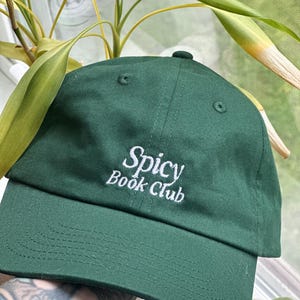 K&ouml;nnte beinhalten: Dunkelgr&uuml;ne Baseballkappe mit den wei&szlig; gestickten Worten "Spicy Book Club". Die Kappe hat einen gebogenen Schirm und einen Knopf oben. Der Hut ist aus weichem Stoff.
