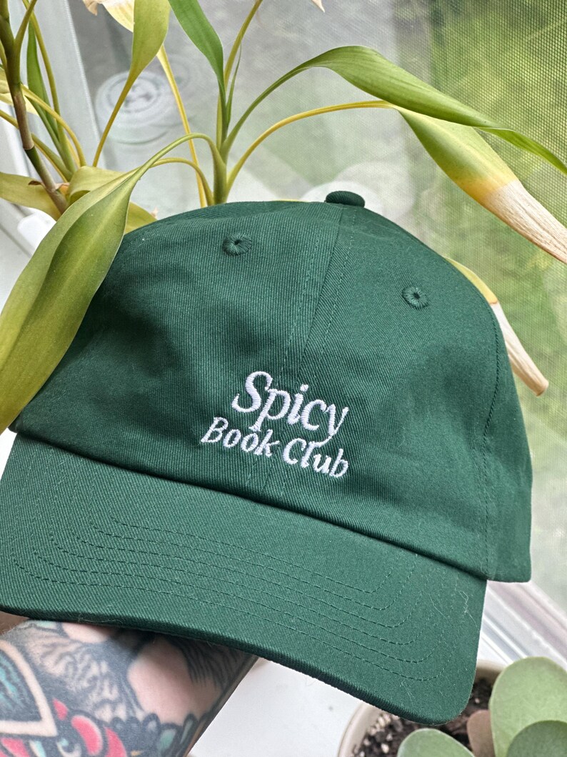 K&ouml;nnte beinhalten: Dunkelgr&uuml;ne Baseballkappe mit den wei&szlig; gestickten Worten "Spicy Book Club". Die Kappe hat einen gebogenen Schirm und einen Knopf oben. Der Hut ist aus weichem Stoff.