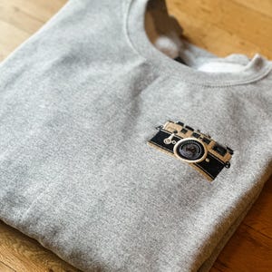 May include: A gray sweatshirt with a crew neck featuring an embroidered camera design on the chest. The camera is black and tan with a circular lens. The sweatshirt is folded on a wooden surface.