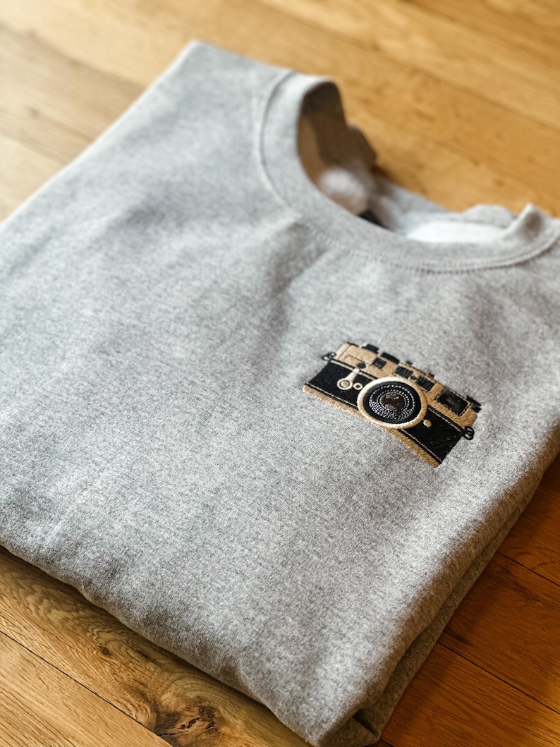 May include: A gray sweatshirt with a crew neck featuring an embroidered camera design on the chest. The camera is black and tan with a circular lens. The sweatshirt is folded on a wooden surface.