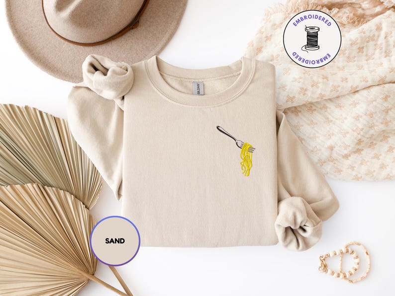 May include: Sand-colored sweatshirt with an embroidered design of a fork holding yellow spaghetti. The sweatshirt has long sleeves and a crew neck. A tan hat and a dried palm leaf are in the background.