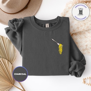May include: Charcoal gray sweatshirt with a small embroidered design of spaghetti on a fork. The sweatshirt has long sleeves and a crew neck. The word "CHARCOAL" is printed on a circular label.