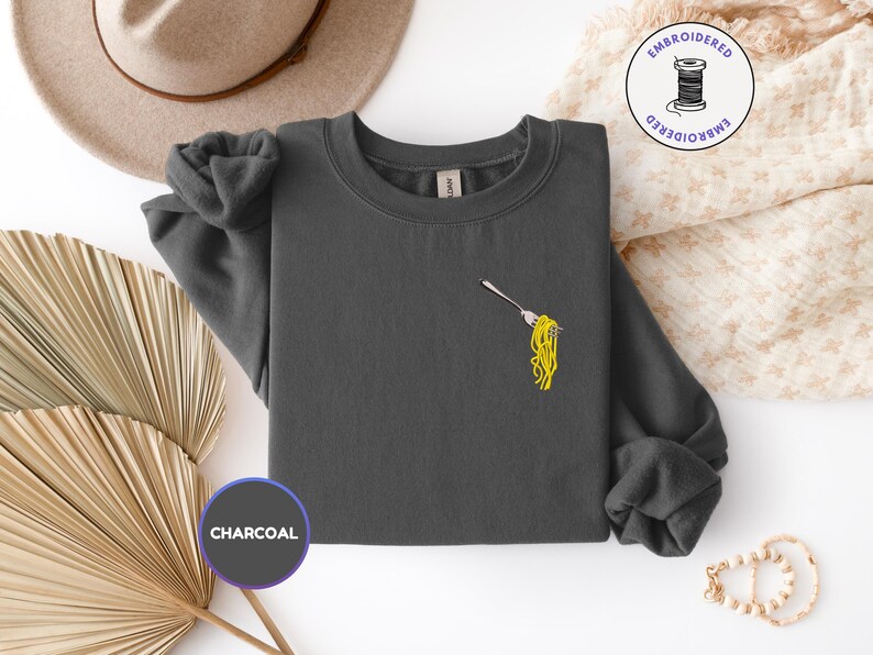 May include: Charcoal gray sweatshirt with a small embroidered design of spaghetti on a fork. The sweatshirt has long sleeves and a crew neck. The word "CHARCOAL" is printed on a circular label.