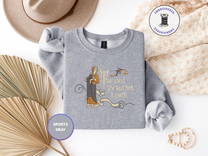 Puede incluir: Una sudadera gris deportivo de cuello redondo con un bordado que muestra una figura de estilo medieval con t&uacute;nica oscura. El texto dice "Thar goes! The last fvck I giveth." El dise&ntilde;o incluye volutas decorativas y un peque&ntilde;o p&aacute;jaro.