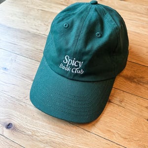 K&ouml;nnte beinhalten: Dunkelgr&uuml;ne Baseballkappe mit dem Text "Spicy Book Club" in Wei&szlig; gestickt. Die Kappe ist aus strapazierf&auml;higem Stoff gefertigt und hat einen gebogenen Schirm. Der Hut liegt auf einer Holzoberfl&auml;che.