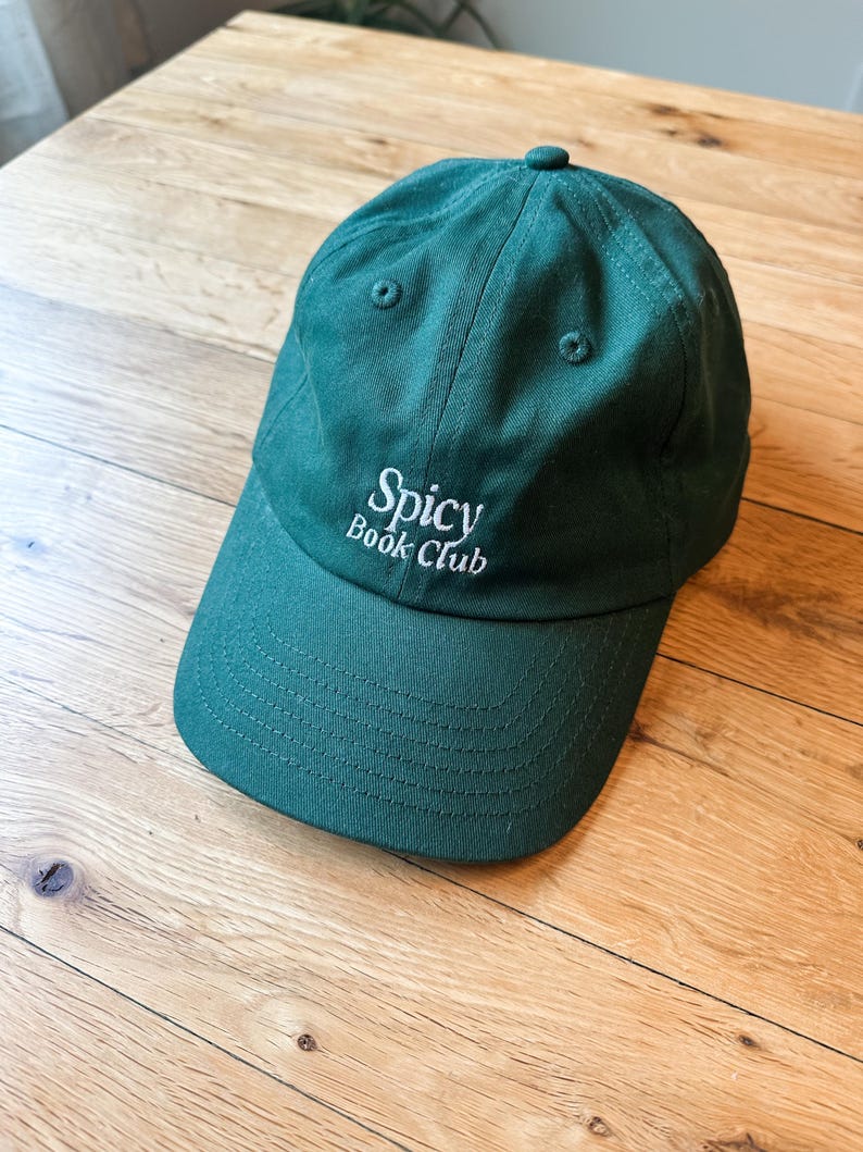 K&ouml;nnte beinhalten: Dunkelgr&uuml;ne Baseballkappe mit dem Text "Spicy Book Club" in Wei&szlig; gestickt. Die Kappe ist aus strapazierf&auml;higem Stoff gefertigt und hat einen gebogenen Schirm. Der Hut liegt auf einer Holzoberfl&auml;che.