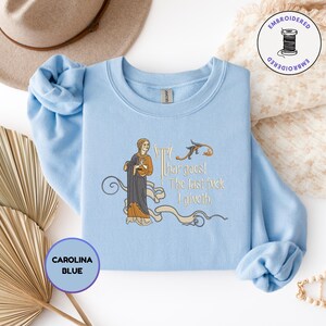 Puede incluir: Una sudadera azul claro de cuello redondo con un bordado de una figura de estilo medieval con t&uacute;nica oscura y detalles dorados. El texto dice "Thar goes! The last fuck I giveth." El dise&ntilde;o incluye adornos de volutas y un peque&ntilde;o p&aacute;jaro.
