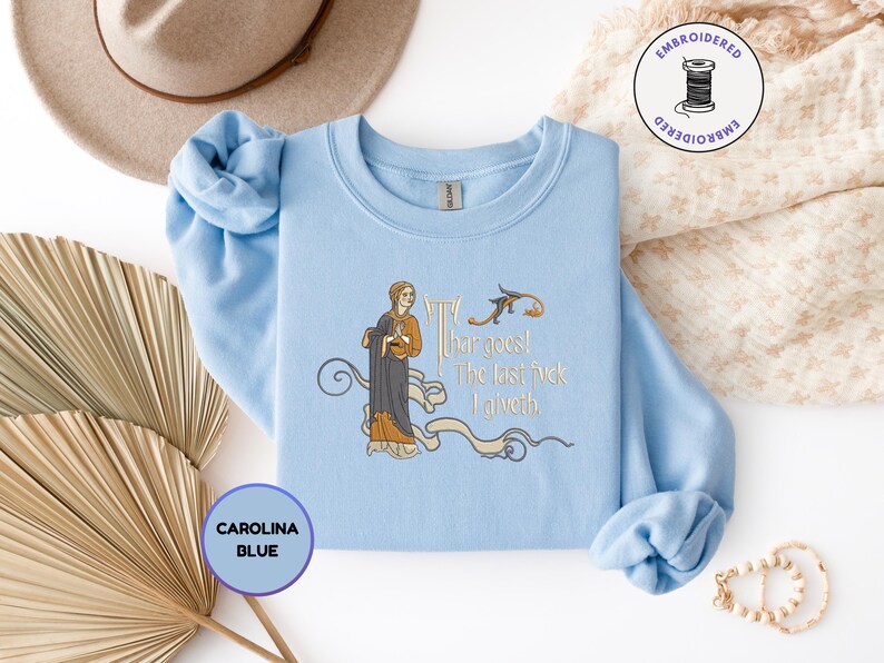 Puede incluir: Una sudadera azul claro de cuello redondo con un bordado de una figura de estilo medieval con t&uacute;nica oscura y detalles dorados. El texto dice "Thar goes! The last fuck I giveth." El dise&ntilde;o incluye adornos de volutas y un peque&ntilde;o p&aacute;jaro.