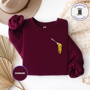 May include: Maroon sweatshirt with an embroidered design of a fork holding yellow spaghetti. The sweatshirt has a crew neck and long sleeves. A hat and decorative items are in the background.