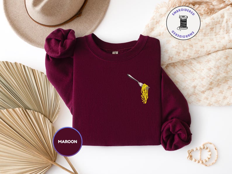 May include: Maroon sweatshirt with an embroidered design of a fork holding yellow spaghetti. The sweatshirt has a crew neck and long sleeves. A hat and decorative items are in the background.
