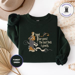 Puede incluir: Una sudadera de cuello redondo color verde bosque con un bordado de estilo medieval que muestra una figura con t&uacute;nica y el texto "Thar goes! The last fvck I giveth". El dise&ntilde;o incluye un peque&ntilde;o drag&oacute;n y adornos decorativos.