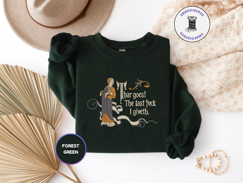 Puede incluir: Una sudadera de cuello redondo color verde bosque con un bordado de estilo medieval que muestra una figura con t&uacute;nica y el texto "Thar goes! The last fvck I giveth". El dise&ntilde;o incluye un peque&ntilde;o drag&oacute;n y adornos decorativos.