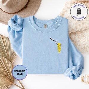 May include: Carolina blue sweatshirt with an embroidered fork holding yellow spaghetti. The sweatshirt has long sleeves and a crew neck. A round sticker with the text "EMBROIDERED" and a spool of thread is in the upper right corner.