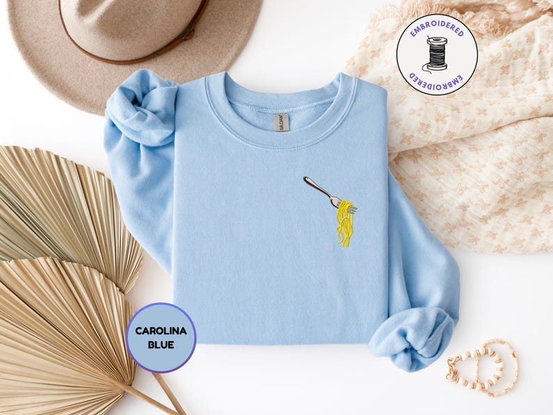 May include: Carolina blue sweatshirt with an embroidered fork holding yellow spaghetti. The sweatshirt has long sleeves and a crew neck. A round sticker with the text "EMBROIDERED" and a spool of thread is in the upper right corner.