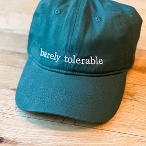 Op de afbeelding: Een donkergroene baseballpet met de geborduurde tekst "barely tolerable" in het wit. De pet heeft een gebogen klep en een knoop aan de bovenkant. De hoed is gemaakt van een zachte stof.