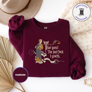 Puede incluir: Una sudadera de cuello redondo color granate con un bordado de estilo medieval que muestra a una persona con t&uacute;nica y el texto: Thar goes! The last fvck I giveth. El dise&ntilde;o incluye adornos en forma de volutas y un peque&ntilde;o p&aacute;jaro en dorado y gris.