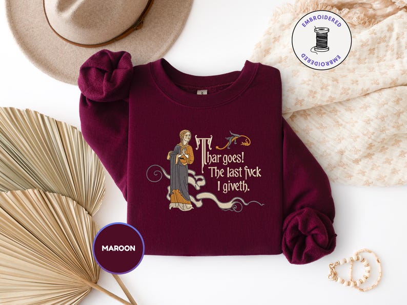 Puede incluir: Una sudadera de cuello redondo color granate con un bordado de estilo medieval que muestra a una persona con t&uacute;nica y el texto: Thar goes! The last fvck I giveth. El dise&ntilde;o incluye adornos en forma de volutas y un peque&ntilde;o p&aacute;jaro en dorado y gris.