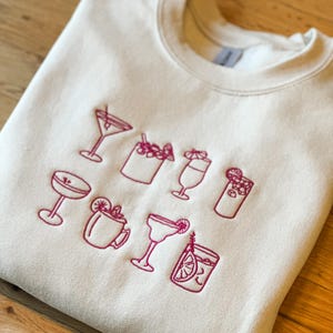 Puede incluir: Una sudadera beige con un diseño bordado en rosa de varios cócteles. El diseño incluye una copa de martini, una copa de margarita, un vaso alto, una coctelera, una taza y un vaso con una rodaja de limón.